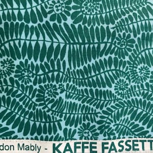 May include: Close-up of fabric with a repeating pattern of dark teal fern-like leaves and spiral floral elements on a light teal background. The text "Brandon Mably - KAFFE FASSETT COL" is visible at the bottom.