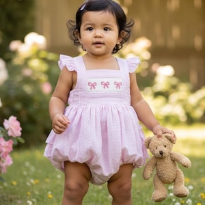 May include: A young child wearing a light pink romper with a bow design and holding a small teddy bear. The romper has a square neckline and ruffled sleeves. The child is standing in a grassy area with flowers in the background.