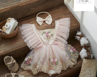 May include: A baby girl's outfit set, including a pink romper with floral embroidery, a matching headband with a bow, and gold-colored baby shoes. The romper features ruffled sleeves and a tulle skirt. A wooden toy and a basket are also in the image.