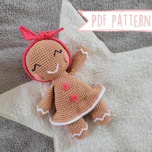May include: Crochet gingerbread person doll with a red bow, white trim, and pink buttons. The doll is lying on a white and gray fuzzy surface. PDF PATTERN FR/ENG