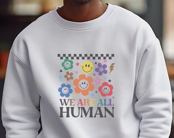 Può includere: Felpa bianca con una grafica colorata di facce sorridenti e fiori. Il testo "WE ARE ALL HUMAN" è stampato sotto la grafica.