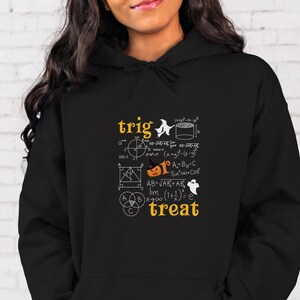 以下が含まれることがあります： 黒のフード付きスウェットシャツに、白でカボチャ、幽霊、魔女のグラフィックプリントが施され、数学の式と「trig treat」の文字が書かれています。