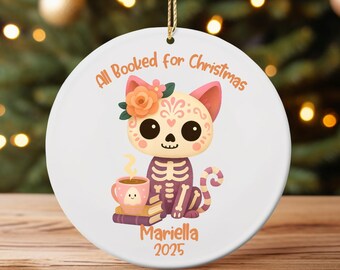 May include: A white ceramic Christmas ornament with a cat skeleton design. The cat has a floral crown and holds a cup of coffee. The text "All Booked for Christmas" and the name "Mariella 2025" are printed on the ornament.