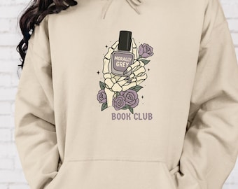 Könnte beinhalten: Beigefarbener Kapuzenpullover mit Grafikdruck. Das Design zeigt eine Skeletthand, die einen Nagellackflakon mit der Aufschrift "Morally Grey" hält. Lila Rosen und "Book Club" sind ebenfalls abgebildet. Der Hoodie hat eine Kapuze mit Kordelzug und eine Vordertasche.