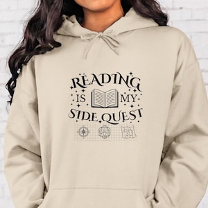 Może przedstawiać: Beżowa bluza z kapturem z napisem "Reading is my side quest" w kolorze czarnym. Tekst jest otoczony gwiazdami i zawiera otwartą książkę, kompas, kostkę do gry i mapę.