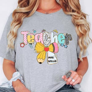 May include: A gray t-shirt with the word "TEACHER" in colorful, patterned letters. The design includes a pencil, apple, and the name "MRS. WILLIS". The shirt has short sleeves.
