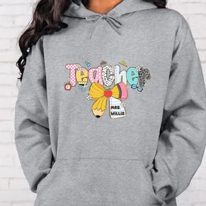 May include: A gray hoodie with a colorful graphic that says "Teacher." The graphic includes a pencil, apple, and bow, with the name "Mrs. Willis" on a tag. The design has a mix of patterns and colors.