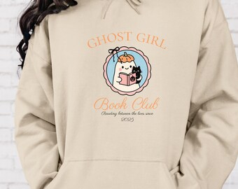 May include: Beige hoodie with a front graphic. The graphic features a cartoon ghost wearing a pumpkin hat, reading a book with a black cat. Text reads "GHOST GIRL Book Club" and "Reading between the lives since 2025".
