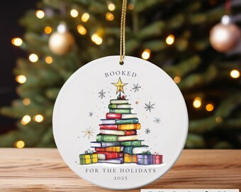 Puede incluir: Adorno navideño redondo de cerámica blanca con una cuerda dorada. El adorno presenta una pila de libros con forma de árbol de Navidad, coronada por una estrella dorada. El texto "BOOKED" y "FOR THE HOLIDAYS 2025" está impreso.