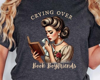 May include: Dark gray t-shirt featuring a vintage illustration of a woman crying while reading a book. The text "Crying Over" is above the illustration, and "Book Boyfriends" is below. The woman is wearing a pearl necklace and has a rose on her dress.