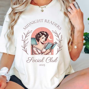May include: White t-shirt with a pink and white illustration of a woman reading a book under a lamp. The text "Midnight Readers Social Club 2023" is printed on the shirt.