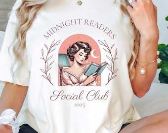Op de afbeelding: Wit T-shirt met een roze en witte illustratie van een vrouw die een boek leest onder een lamp. De tekst "Midnight Readers Social Club 2023" is op het T-shirt gedrukt.