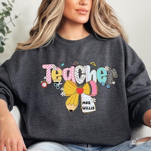 May include: Dark gray sweatshirt with the word "Teacher" in a colorful, patterned design. The design includes a pencil, bow, and the name "Mrs. Willis" on a notepad. The sweatshirt is a comfortable, casual style.