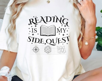May include: White t-shirt with a black graphic that says "Reading is my side quest" with a book, compass, dice, and map illustrations.