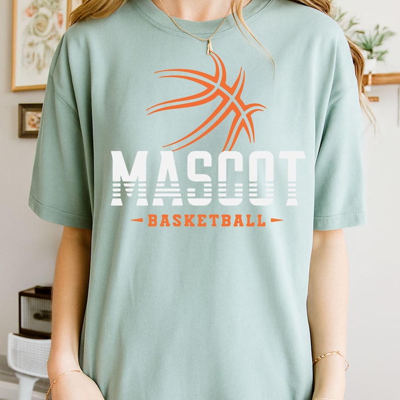 May include: A light green t-shirt with the word "MASCOT" in white block letters and an orange basketball graphic. Below the word "MASCOT" is the word "BASKETBALL" in orange. The t-shirt is a casual style.