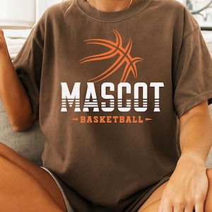 May include: A brown t-shirt featuring a basketball graphic in orange and white. The word "MASCOT" is printed in white block letters, with "BASKETBALL" below in orange. The shirt has short sleeves and a crew neck.
