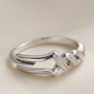 May include: A silver-toned ring featuring a polished, open-work design with a smooth, angular band that transitions into a textured, twisted section on one side. The metal surface reflects light, highlighting the intricate, layered structure of the band.
