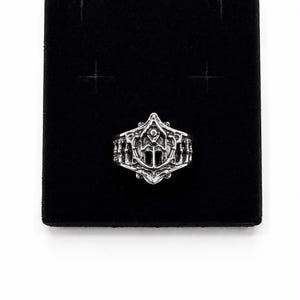 May include: A silver-toned metal ring featuring an intricate, raised relief design with symmetrical, ornate patterns and a central pointed motif. The band displays a textured, sculpted surface with deep grooves and a polished metallic finish.