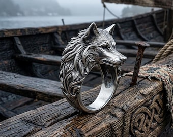 May include: A silver ring featuring a detailed wolf head design. The ring is set against a backdrop of a wooden boat, with a blurred background of water and a foggy sky. The wolf's fur and facial features are intricately carved.