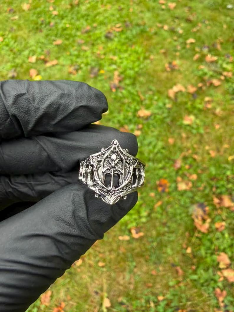 May include: A silver-toned metal ring featuring an intricate, raised relief design with a central pointed shield-like motif and symmetrical ornate carvings along the band, showcasing a polished finish with darkened recessed areas for contrast.