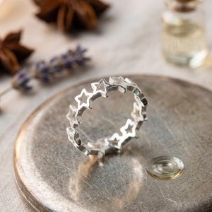 May include: A silver ring featuring a repeating star design. The ring is set on a silver-colored surface, with a small glass bottle and dried botanicals in the background. The ring's design is a series of open star shapes.