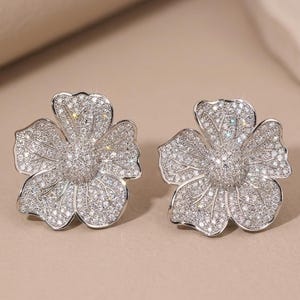 May include: A pair of silver-toned stud earrings shaped like five-petaled flowers. Each earring is densely encrusted with numerous small, clear, sparkling stones that cover the entire surface of the petals and the raised, rounded center.