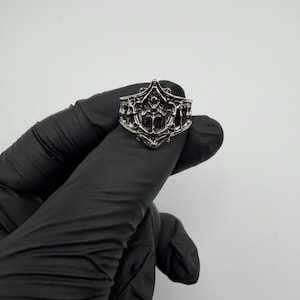 May include: A silver-toned metal ring featuring an intricate, openwork design with raised filigree patterns and a pointed, crown-like top edge. The band has a dark, oxidized finish in the recessed areas, highlighting the detailed, symmetrical metalwork.