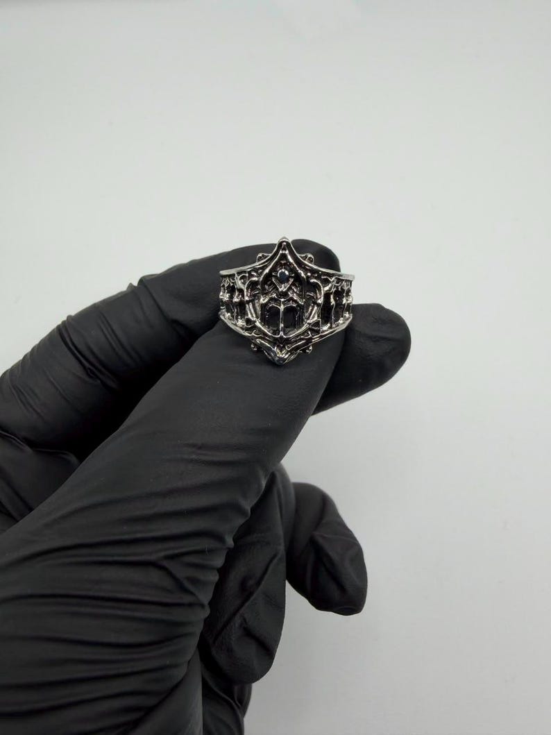 May include: A silver-toned metal ring featuring an intricate, openwork design with raised filigree patterns and a pointed, crown-like top edge. The band has a dark, oxidized finish in the recessed areas, highlighting the detailed, symmetrical metalwork.