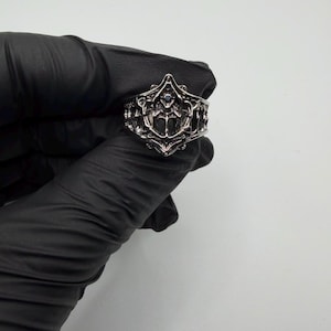 May include: A silver-toned metal ring featuring an intricate, openwork design with raised, ornate filigree patterns and a small, dark, circular stone set centrally within the detailed, pointed, and symmetrical decorative face of the band.