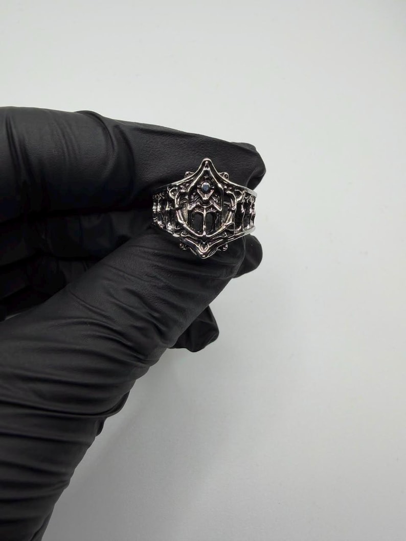 May include: A silver-toned metal ring featuring an intricate, openwork design with raised, ornate filigree patterns and a small, dark, circular stone set centrally within the detailed, pointed, and symmetrical decorative face of the band.