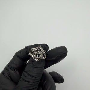 May include: A silver-toned metal ring featuring an intricate, openwork design with a central pointed arch motif and vertical bar detailing. The band has a textured, oxidized finish that highlights the detailed relief patterns across the surface.