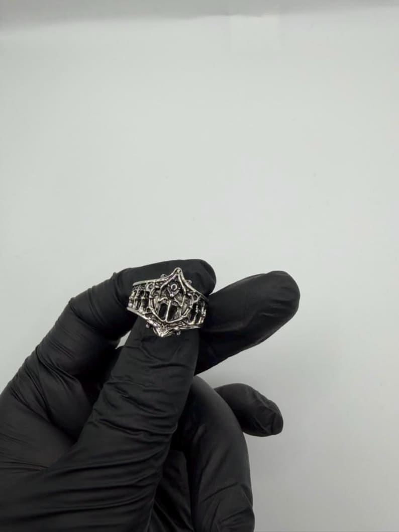 May include: A silver-toned metal ring featuring an intricate, openwork design with a central pointed arch motif and vertical bar detailing. The band has a textured, oxidized finish that highlights the detailed relief patterns across the surface.