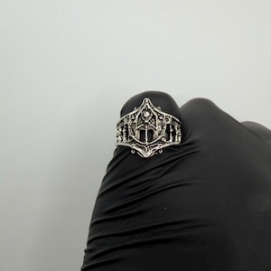 May include: A silver-toned metal ring featuring an intricate, openwork design with a pointed, arch-like centerpiece. The band displays a textured, filigree-style pattern with symmetrical cutouts and a small, embedded clear stone at the top peak.