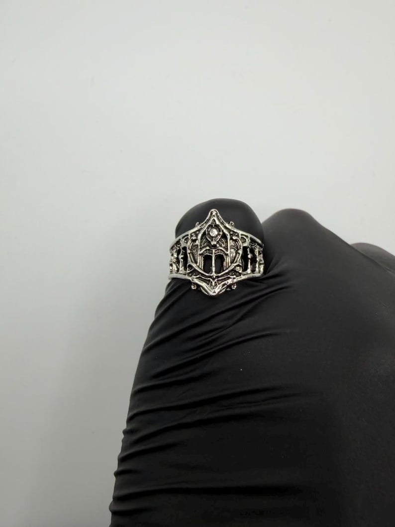 May include: A silver-toned metal ring featuring an intricate, openwork design with a pointed, arch-like centerpiece. The band displays a textured, filigree-style pattern with symmetrical cutouts and a small, embedded clear stone at the top peak.