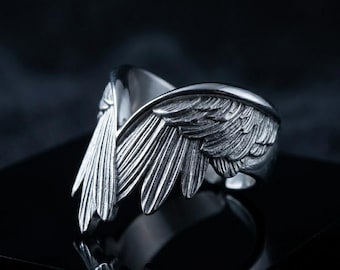 May include: A silver-toned metal ring featuring a detailed design of two symmetrical wings that meet at the center. The surface of the wings displays intricate, etched feather textures with darkened recessed areas to highlight the individual plumage.