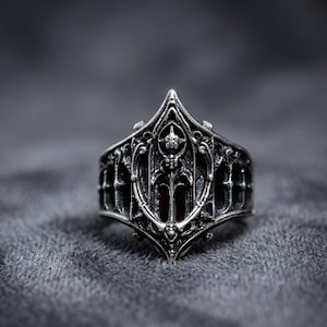 May include: A silver-toned metal ring featuring an intricate, pointed architectural design with arched cutouts and a central shield-like motif. The metal has a dark, oxidized finish in the recessed areas, highlighting the detailed relief work.