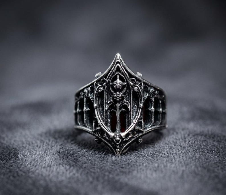 May include: A silver-toned metal ring featuring an intricate, pointed architectural design with arched cutouts and a central shield-like motif. The metal has a dark, oxidized finish in the recessed areas, highlighting the detailed relief work.