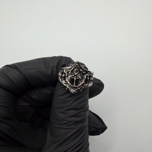 May include: A silver-toned metal ring featuring an intricate, raised relief design with pointed, arch-like structures and a central vertical element. The band has a textured, darkened finish within the recessed areas to highlight the detailed metalwork.