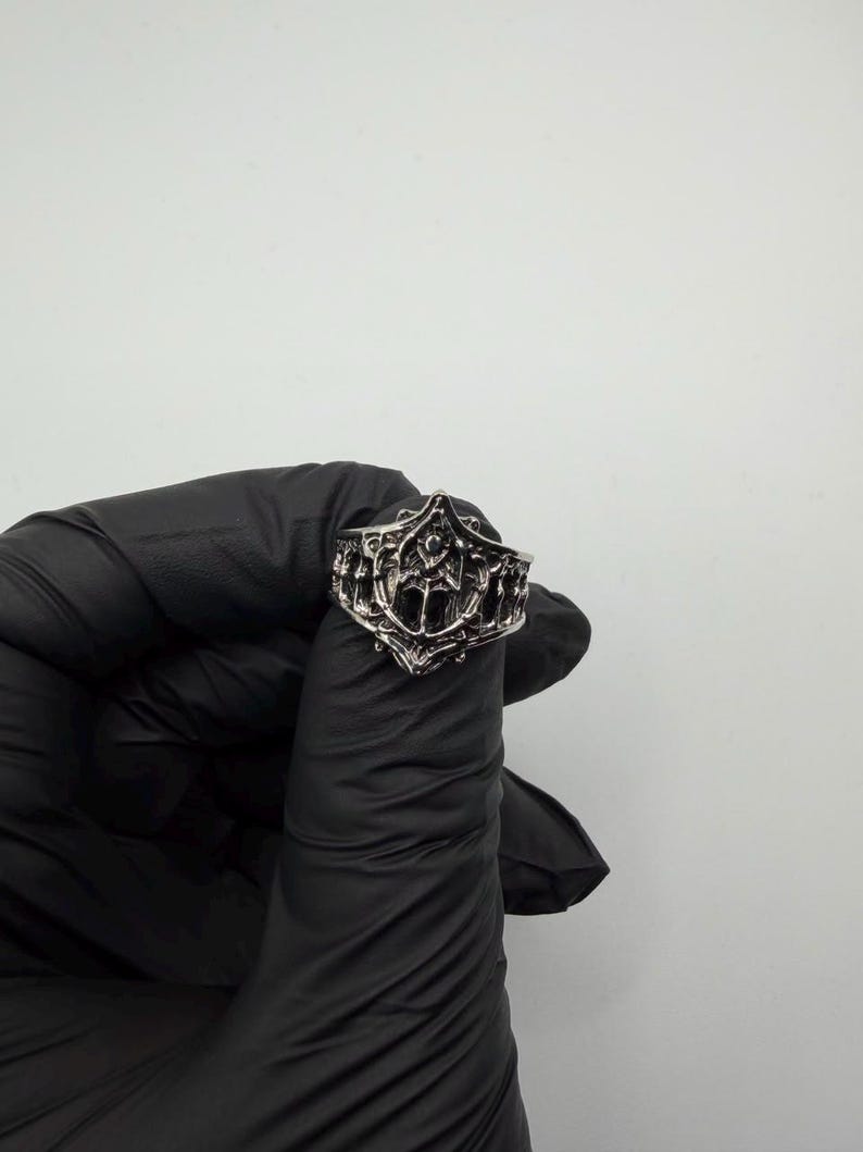 May include: A silver-toned metal ring featuring an intricate, raised relief design with pointed, arch-like structures and a central vertical element. The band has a textured, darkened finish within the recessed areas to highlight the detailed metalwork.