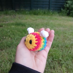 Könnte beinhalten: Eine handgefertigte, farbenfrohe Häkelbiene mit Regenbogenstreifen, gelbem Körper und weißen Flügeln. Das Plüschtier wird in einer Hand vor dem Hintergrund von grünem Gras und einem Holzzaun gehalten.