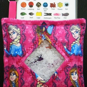 Puede incluir: Juguete sensorial rosa con una ventana transparente llena de cuentas blancas. El juguete est&aacute; decorado con im&aacute;genes de los personajes de la pel&iacute;cula Frozen. El juguete tiene una etiqueta con las palabras "Puzzle Piece", "Soccer Ball", "Star", "Heart", "Lion", "Bear", "Ring", "Penny", "Ladybug", "Bell", "Smiley Ball", "Train", "Flower", "Eye", "Leaf", "Shell", "Lego", "Clothespin", "Flip Flop", and "Fish".