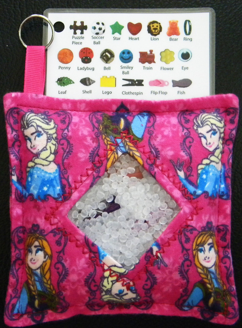 Puede incluir: Juguete sensorial rosa con una ventana transparente llena de cuentas blancas. El juguete est&aacute; decorado con im&aacute;genes de los personajes de la pel&iacute;cula Frozen. El juguete tiene una etiqueta con las palabras "Puzzle Piece", "Soccer Ball", "Star", "Heart", "Lion", "Bear", "Ring", "Penny", "Ladybug", "Bell", "Smiley Ball", "Train", "Flower", "Eye", "Leaf", "Shell", "Lego", "Clothespin", "Flip Flop", and "Fish".