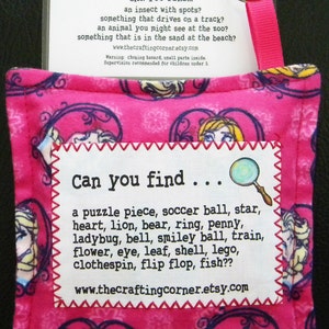 May include: Pink fabric pouch with a white tag that says "Can you find..." and lists a variety of items. The pouch has a pink ribbon attached to it and a clear plastic card with the text "The Crafting Corner" and "Can you find..." with a list of items.