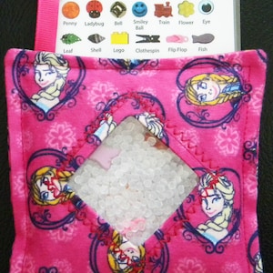 May include: Pink fabric sensory bag with a clear window filled with white beads. The bag has a Frozen theme with images of Elsa and Anna. The bag has a zipper closure and a loop for hanging.