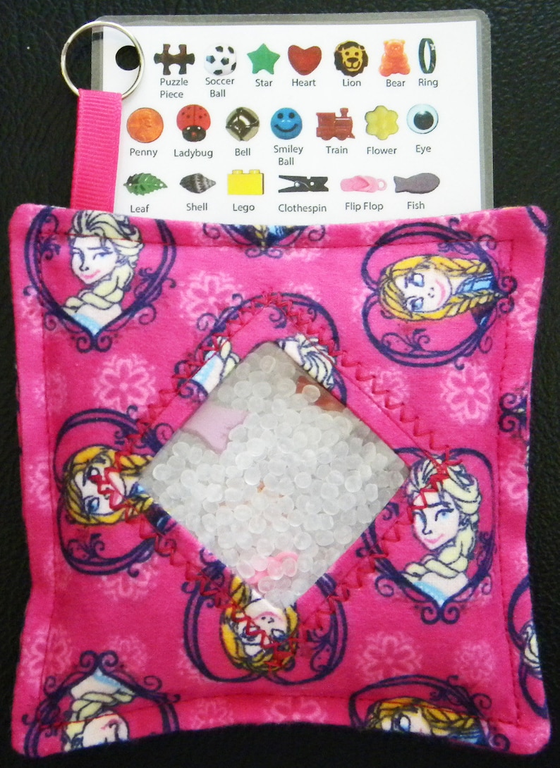 May include: Pink fabric sensory bag with a clear window filled with white beads. The bag has a Frozen theme with images of Elsa and Anna. The bag has a zipper closure and a loop for hanging.