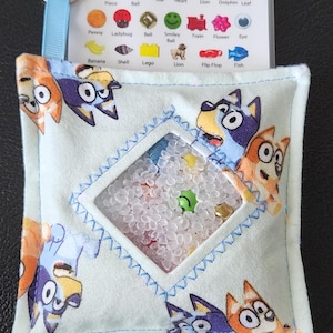 May include: A blue and white sensory toy with a clear window filled with small beads. The toy has a zippered pouch and a tag with images of various objects, including a puzzle piece, basket ball, star, heart, lion, dolphin, leaf, penny, ladybug, bell, smiley face, train, flower, eye, banana, shell, lego, lion, flip flop, and fish.