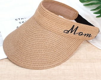 May include: A tan straw visor featuring a wide, curved brim and a structured headband. The word Mom is embroidered in black cursive script on the front of the headband. A black elastic strap is attached to the back for an adjustable fit.