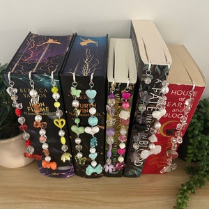 Puede incluir: Seis sujetalibros con abalorios de cuentas de colores unidos a clips plateados. Los abalorios incluyen mariposas, corazones, flores y estrellas. Los sujetalibros están unidos a libros con títulos como "A Fate Inked In Blood", "House of Earth and Blood" y "In the Flame I Rise".