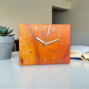 May include: A wooden desk clock with a square face and a warm orange hue. The clock has silver hands and markers, and is sitting on a white surface. A succulent plant in a gray pot and an open book are also visible.