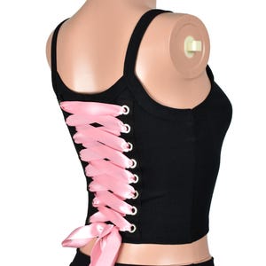 May include: Black crop top with a pink ribbon lace-up back. The top has a scoop neckline and thin straps. The pink ribbon is laced through silver eyelets. The top is paired with a black skirt.